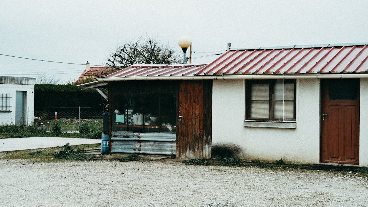 A rustic and shabby shed with a red metal roof, set outdoors in a rural area.