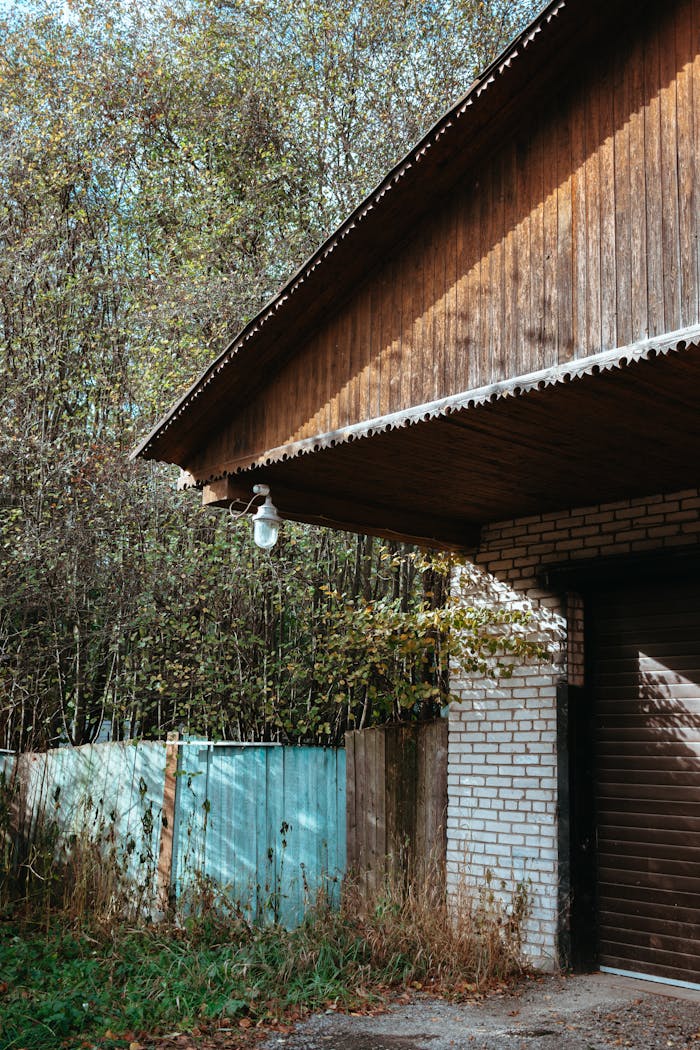 A serene rustic garage with a wooden roof and brick walls next to a fence amidst trees.